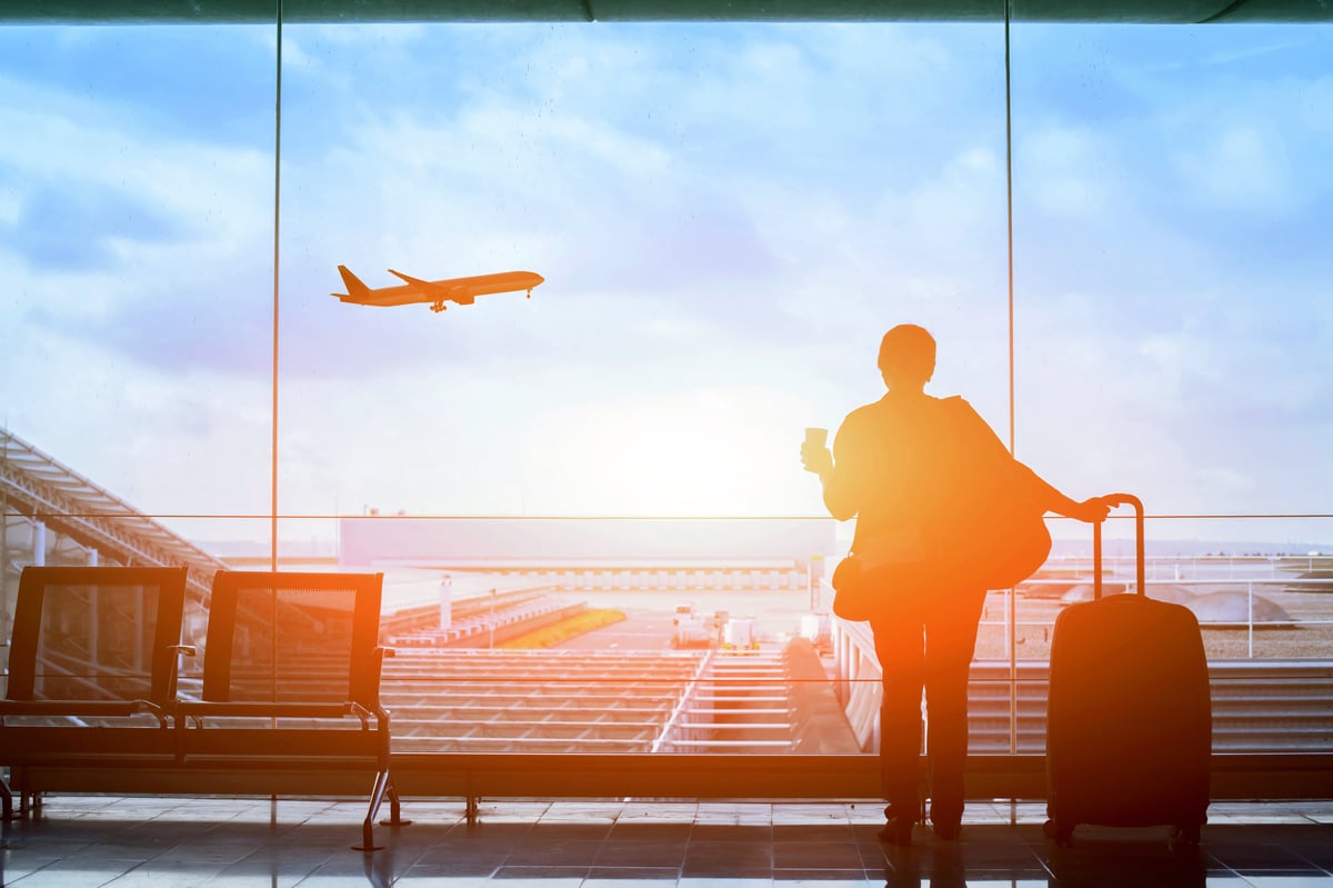 Image of a woman standing in an airport, in front of a large window with the sunset and planes landing outside.